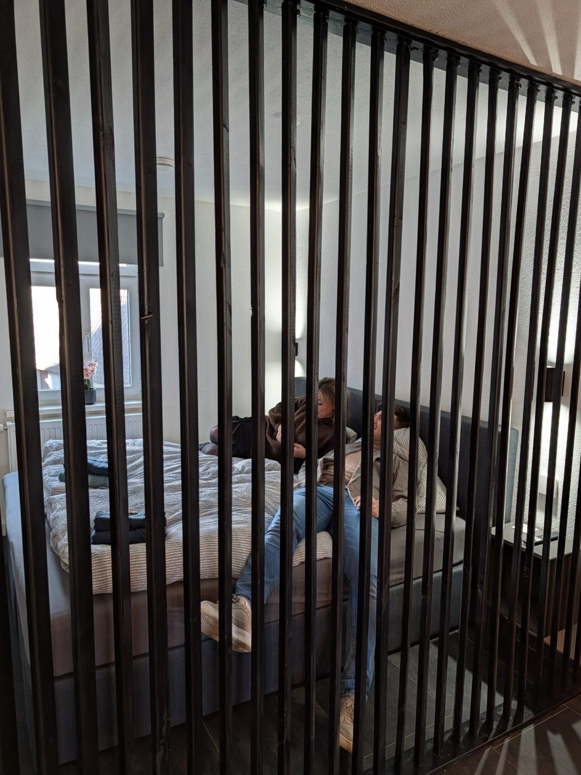 Zwei Personen sitzen entspannt auf einem Bett, von senkrechten Streifen verdeckt.