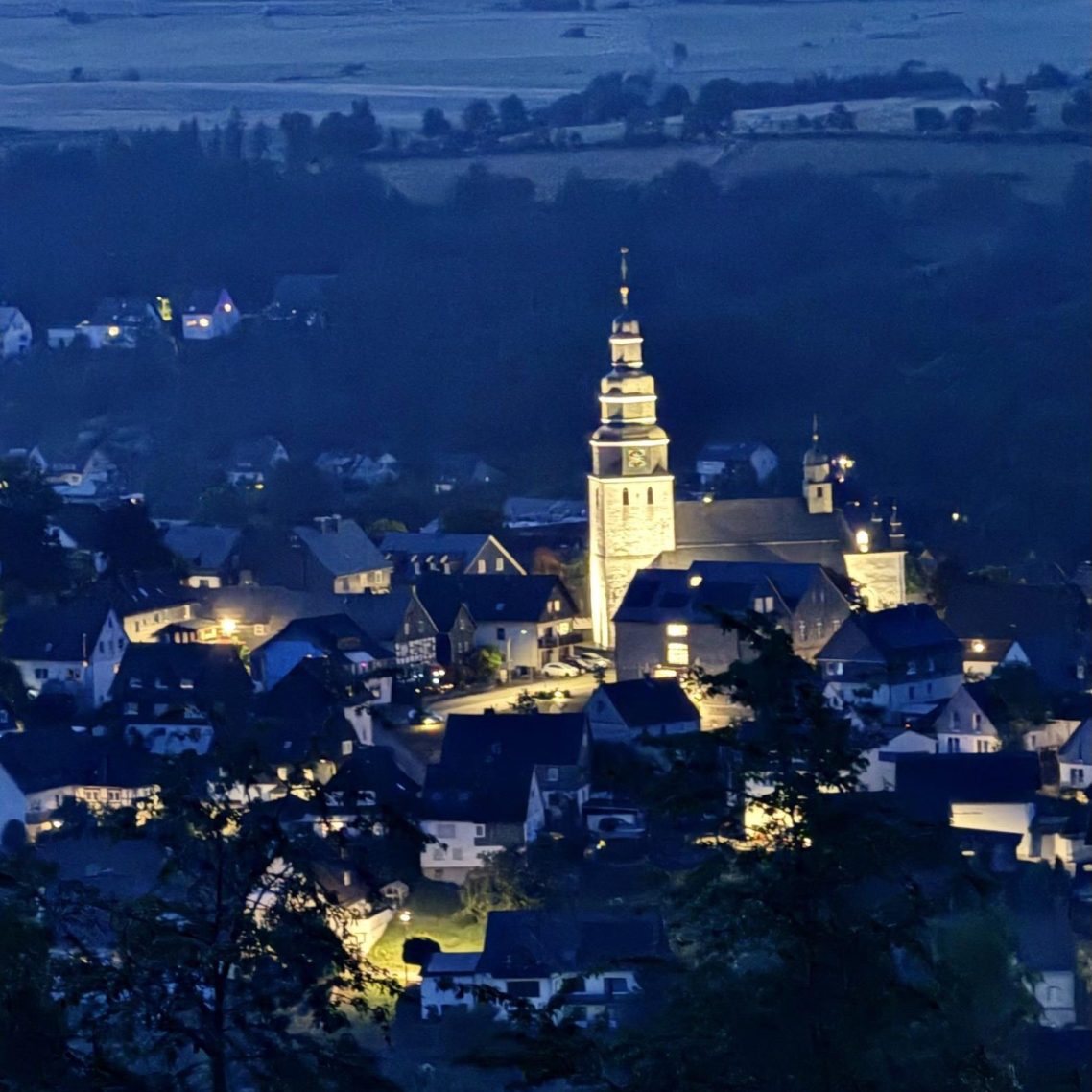 Nachtaufnahme eines beleuchteten Dorfes mit Kirche in ländlicher Umgebung.